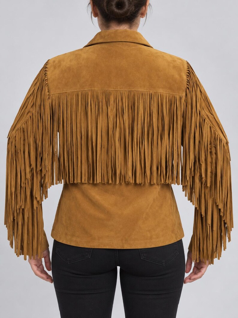 K&ouml;nnte beinhalten: Eine hellbraune Wildlederjacke mit langen Fransen am R&uuml;cken und an den &Auml;rmeln. Die Jacke hat einen klassischen Kragen und einen geraden Saum, was ihr durch die weiche, matte Oberfl&auml;che und die einheitliche erdige Farbe eine Textur verleiht.