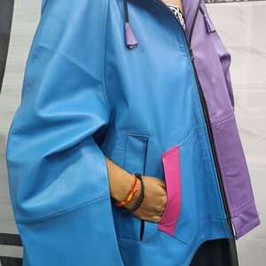 May include: A color-blocked hooded jacket featuring a bright blue main body, a purple left panel, and a vibrant pink pocket trim. The jacket includes a front zipper closure, a hood with a purple interior lining, and wide, cape-style sleeves.