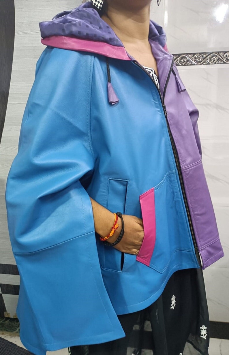 May include: A color-blocked hooded jacket featuring a bright blue main body, a purple left panel, and a vibrant pink pocket trim. The jacket includes a front zipper closure, a hood with a purple interior lining, and wide, cape-style sleeves.