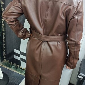 May include: A long, brown leather trench coat featuring a wide collar, long sleeves, and a matching belt cinched at the waist. The coat has a smooth finish, a central back vent, and structured shoulder detailing, extending down to the lower calf area.