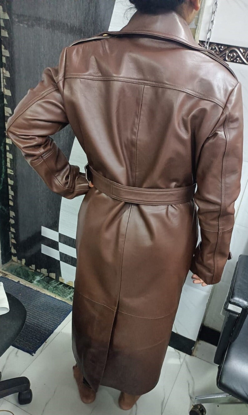 May include: A long, brown leather trench coat featuring a wide collar, long sleeves, and a matching belt cinched at the waist. The coat has a smooth finish, a central back vent, and structured shoulder detailing, extending down to the lower calf area.