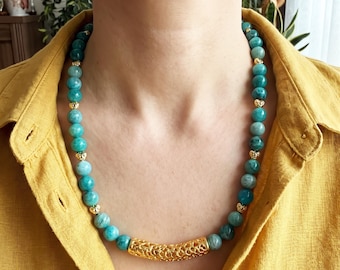 May include: A beaded necklace featuring round, teal-coloured stones interspersed with small, gold-toned metal spacers. The necklace includes a central, elongated, gold-toned cylindrical bead with an intricate, open-work filigree pattern.