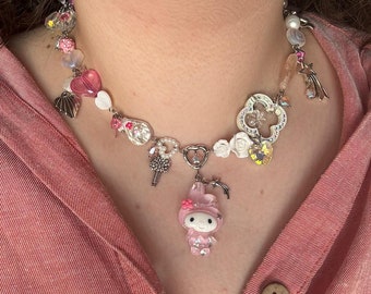 May include: A whimsical necklace featuring a variety of charms and beads. The necklace includes pink heart-shaped beads, white flower accents, and a pendant of a pink cartoon character. Additional charms include a key, a star, and a shell.