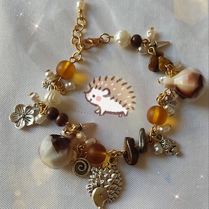 Puede incluir: Un brazalete de oro con una variedad de dijes, incluyendo un erizo, una flor, un remolino y una concha. El brazalete también presenta cuentas marrones y blancas, y una cadena de oro.