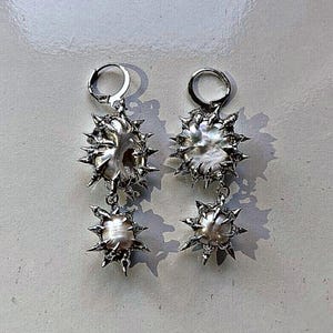 Pode incluir: Um par de brincos prateados com um design único. Cada brinco apresenta uma pérola grande e irregular, rodeada por uma moldura metálica pontiaguda em forma de estrela. Uma pérola menor pende por baixo, também envolvida numa moldura pontiaguda.