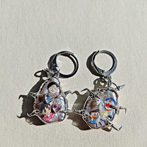 Può includere: Orecchini in argento con forme irregolari a forma di perla, ciascuno decorato con personaggi dei cartoni animati. Gli orecchini hanno una montatura metallica appuntita e una chiusura a leva. I personaggi dei cartoni animati sono dai colori vivaci.