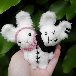 Puede incluir: Dos muñecos de peluche de conejito blanco con detalles cosidos en negro y ojos negros. Un conejito lleva un collar rosa, el otro un collar negro. Los conejitos se sostienen en una mano sobre un fondo de hojas verdes.