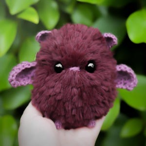 Puede incluir: Una criatura de peluche hecha a mano, de color burdeos, con un cuerpo esponjoso y pequeñas orejas y alas moradas de ganchillo. Tiene ojos grandes, negros y brillantes. El fondo es un follaje verde borroso.