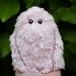 Könnte beinhalten: Ein weiches, plüschiges, lavendelfarbenes Wesen mit einer flauschigen, strukturierten Oberfläche und zwei schwarzen, perlenartigen Augen. Das handgefertigte Spielzeug befindet sich vor einem Hintergrund aus grünem Laub.