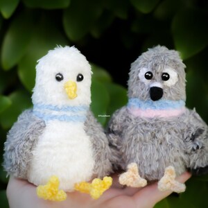 May include: Two plush pigeon toys, one white and gray with a blue collar, the other gray with a pink and blue collar. Both have yellow feet and black eyes. The background is a blurred green foliage.