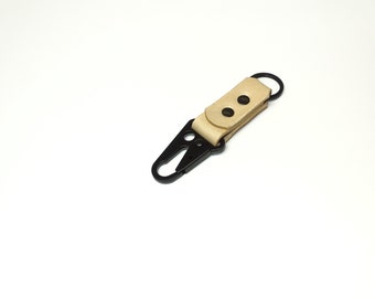 Può includere: Un portachiavi in ​​cuoio beige con un moschettone in metallo nero e due rivetti neri.