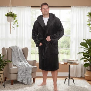 May include: A black hooded robe with a matching belt and pockets. The plush robe is knee-length and is being worn indoors. The person is standing in front of a window with a chair and side table visible.