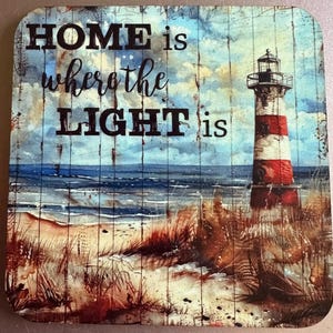 May include: Square coaster with a coastal design. The image features a lighthouse with red and white stripes, a sandy beach, and the ocean under a cloudy sky. Text reads "HOME is where the LIGHT is".