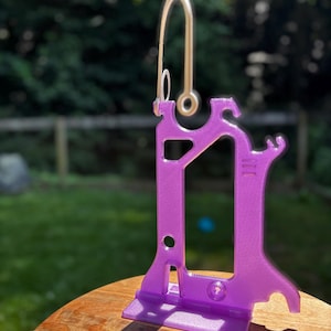 Translucent purple Hero Tubie™ pump stand for the Kangaroo™ Omni, shown outdoors on a wooden surface. The removable, flexible hanger is detached and standing upright, highlighting its curved hook shape for versatile hanging options. The stand’s lightweight 3D-printed structure and fine texture are visible in the sunlight, with a blurred green yard in the background.