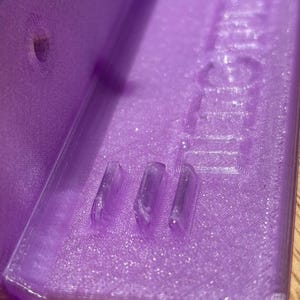 Close-up of a translucent purple 3D-printed plastic base from the Hero Tubie™ system, showing three small integrated line clips designed to prevent kinks in feeding pump tubing. The textured surface and fine print lines of the 3D print are clearly visible, along with a mounting hole on the left. The base is resting on a wooden surface under bright lighting.