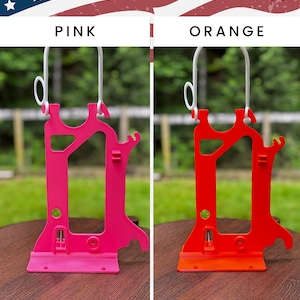 Side-by-side comparison of two Hero Tubie™ pump stands for the Kangaroo™ Omni, under an American flag-themed banner. The left stand is a bright “Pink,” while the right stand is a vivid “Orange.” Both are lightweight, 3D-printed designs with a detachable white hanger for holding a single feeding bag. They’re displayed on wooden tabletops outdoors, with blurred green grass and fencing in the background. Text at the bottom reads “Kangaroo OMNI Single Bag Hero Tubie.”