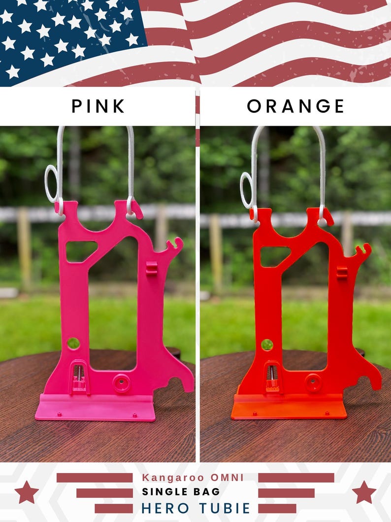 Side-by-side comparison of two Hero Tubie™ pump stands for the Kangaroo™ Omni, under an American flag-themed banner. The left stand is a bright “Pink,” while the right stand is a vivid “Orange.” Both are lightweight, 3D-printed designs with a detachable white hanger for holding a single feeding bag. They’re displayed on wooden tabletops outdoors, with blurred green grass and fencing in the background. Text at the bottom reads “Kangaroo OMNI Single Bag Hero Tubie.”