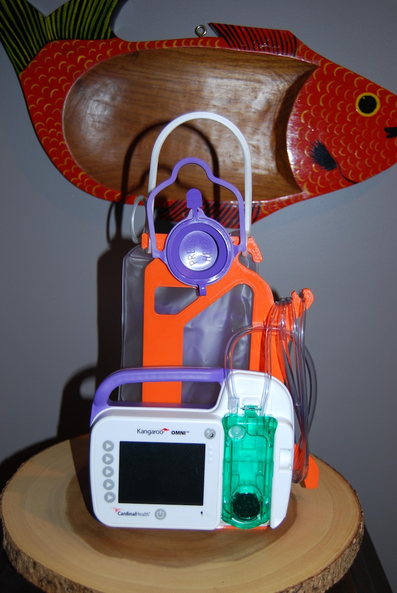 A Kangaroo™ OMNI feeding pump is securely mounted to a bright orange 3D-printed Hero Tubie™ stand. The setup includes a 500ml single enteral feeding bag attached with a flexible and detachable white hanger. The tubing is neatly arranged using included clips to prevent kinks. The unit sits on a wooden platform, with a red fish wall decor in the background. This photo showcases the portable and compact design of the Hero Tubie™ system for G-tube users.