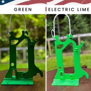 Side-by-side comparison of two Hero Tubie™ pump stands for the Kangaroo™ Omni, shown under an American flag-themed banner. The left stand is a darker “Green” color, while the right is a bright “Electric Lime.” Both stands are lightweight, 3D-printed plastic designs with a detachable white hanger for hanging a single feeding bag. They are displayed on wooden surfaces outdoors with blurred greenery in the background. Text at the bottom reads “Kangaroo OMNI Single Bag Hero Tubie.”
