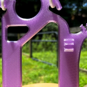 Close-up of the translucent purple 3D-printed Hero Tubie™ hanger piece, shown outdoors with sunlight highlighting the texture and print lines. The design features a circular cutout for hanging the Kangaroo™ Omni pump bag, angled support arms, and integrated line clips on the side to prevent tubing kinks. A green lawn and black fence are visible in the blurred background.
