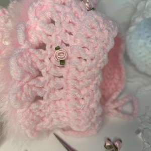 May include: A handmade, pale pink crochet bonnet with a delicate floral design. The bonnet features a soft, fluffy trim and is adorned with small, pink fabric roses. A charming accessory for babies.