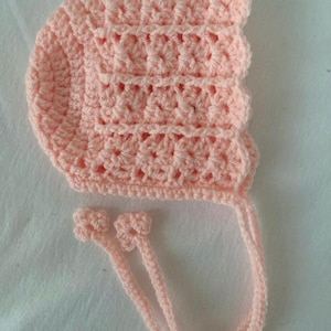Puede incluir: Un gorro de bebé de ganchillo rosa claro. El gorro presenta un patrón texturizado y se ata con dos delicadas cuerdas a juego. El gorro es de un color pastel suave y el ganchillo parece hecho a mano.