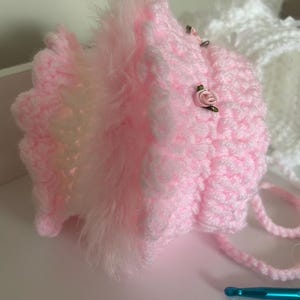 May include: A handmade pink crochet bonnet with a fluffy trim and small rose embellishments. The bonnet has a textured stitch and a tie closure. A white crochet bonnet is visible in the background.