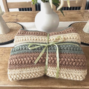 Peut inclure: Une couverture pliée, faite à la main au crochet, dans des tons beige, marron et gris, nouée avec un ruban vert clair. La couverture présente un motif de point texturé et est présentée sur une surface en bois.