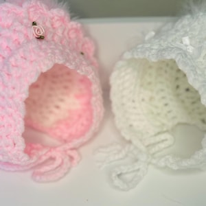 May include: Two handmade baby bonnets. One is light pink with small rose accents and the other is white. Both bonnets are crocheted with a scalloped brim and feather trim. The bonnets have ties.