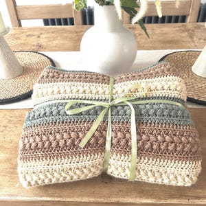 May include: A folded rectangular crochet blanket featuring horizontal stripes in shades of cream, tan, and sage green. The textile displays a variety of textured stitch patterns, including bobble and cable stitches, and is tied with a light green ribbon.
