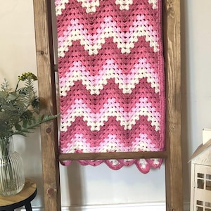 Op de afbeelding: Een handgehaakte deken met een herhalend chevronpatroon in tinten donkerroze, lichtroze en crème. Het textiel heeft een geschulpte randafwerking en is gedrapeerd over een houten ladder, wat het ingewikkelde garenwerk en de textuur laat zien.