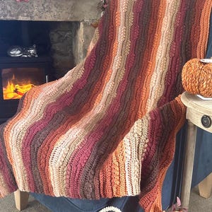 May include: A crochet blanket featuring vertical stripes in shades of dark brown, burgundy, rust orange, and cream. The textile displays a textured pattern with alternating cable-like stitches and rows of raised bobbles throughout the entire design.