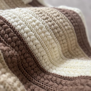 Peut inclure: Une couverture crochetée présentant des rayures verticales alternées de couleur crème, beige et marron. Le textile affiche une variété de points texturés, incluant des points noisette en relief, des rangs plats et des motifs de torsades.