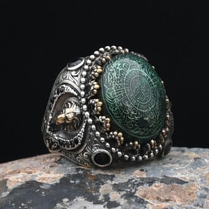 Puede incluir: Un anillo de plata con una piedra verde engastada en un diseño detallado. La piedra tiene una intrincada escritura blanca.