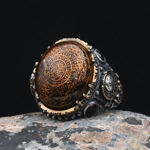 Puede incluir: Un anillo de plata con una piedra grande, redonda y naranja que presenta una intrincada caligrafía árabe. El anillo está decorado con piedras más pequeñas de color negro y blanco.