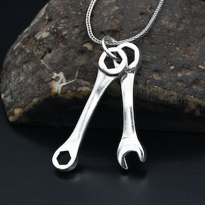 May include: A silver necklace with a pendant featuring two wrenches intertwined. The necklace has a silver chain.