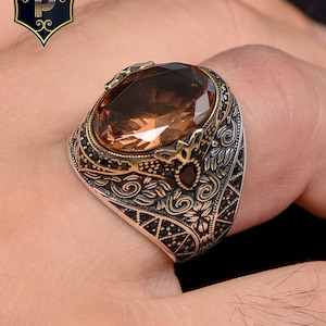 Può includere: Un anello d'argento con un designo ornato e una grande pietra preziosa ovale di colore marrone.