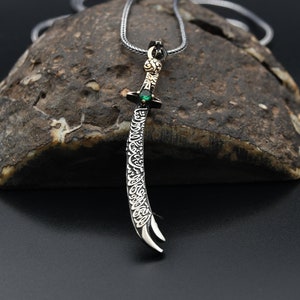May include: A silver pendant necklace with a curved sword design. The sword features detailed engraving and a green gemstone. The pendant hangs from a silver chain, set against a textured stone background.