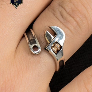May include: Silver-toned adjustable wrench ring with a detailed design. The ring features a realistic wrench head and a band that wraps around the finger. The ring is a unique piece of jewelry.