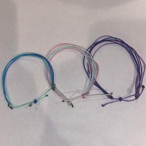 May include: Three colorful friendship bracelets, one blue and green, one pink and white, and one purple. The bracelets are made of woven string and have a knotted closure.