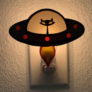 May include: A black and white nightlight shaped like a flying saucer with a cat silhouette in the center. The nightlight is plugged into a standard electrical outlet.