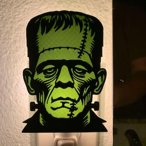 May include: Green Frankenstein's monster night light plug-in. The night light is made of plastic and has a black outline. The monster's face is green and has stitches on the forehead and chin.