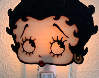 May include: A white night light plug-in with a black and white cartoon image of a woman with a red mouth and large eyes. The night light is lit and glowing orange.