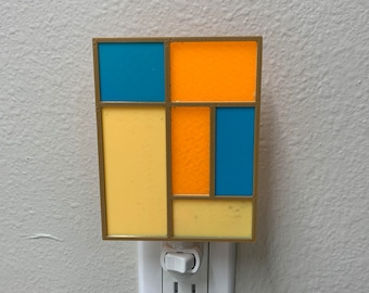May include: A night light with a geometric design featuring yellow, orange, and blue squares. The night light is plugged into an electrical outlet.