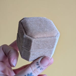 Puede incluir: Una caja hexagonal beige para anillos hecha de material de terciopelo suave. La caja está abierta, revelando el interior. La caja se sostiene en una mano sobre un fondo amarillo. La caja tiene una textura acanalada.