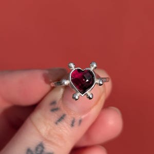 May include: A silver ring with a heart-shaped, deep red gemstone. The ring has a delicate band and small, round accents around the heart. The background is a solid, vibrant red.