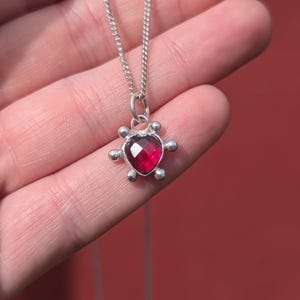 Puede incluir: Un collar de plata con un colgante de piedra roja en forma de corazón. El colgante está rodeado de pequeñas cuentas plateadas. El collar se muestra sobre un fondo rojo.