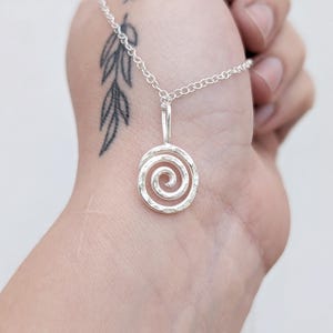 May include: A silver pendant necklace featuring a spiral design. The pendant is suspended from a delicate chain. The spiral has a textured surface. The necklace is shown against a light background.