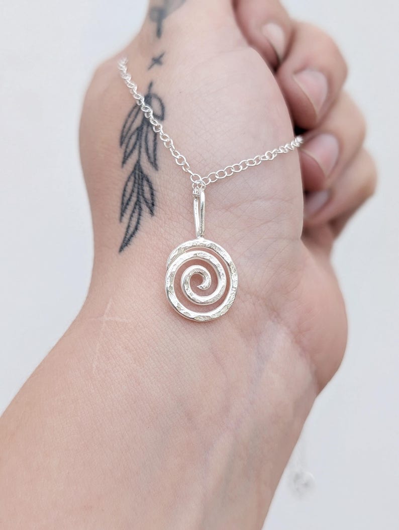 May include: A silver pendant necklace featuring a spiral design. The pendant is suspended from a delicate chain. The spiral has a textured surface. The necklace is shown against a light background.