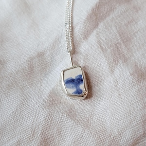 May include: A silver necklace featuring a rectangular pendant with a white background and a blue abstract design. The pendant is encased in a silver frame and hangs from a delicate silver chain. The necklace is displayed on a white linen surface.
